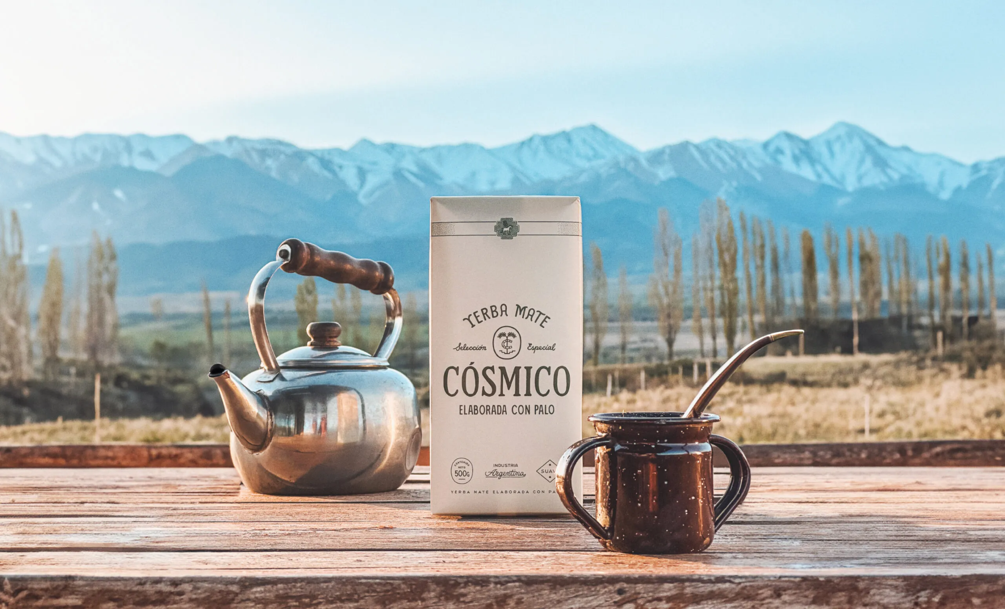 Paquete de Yerba Mate Cósmico sobre una mesa de madera, junto a tetera y mate con bombilla, con montañas de fondo
